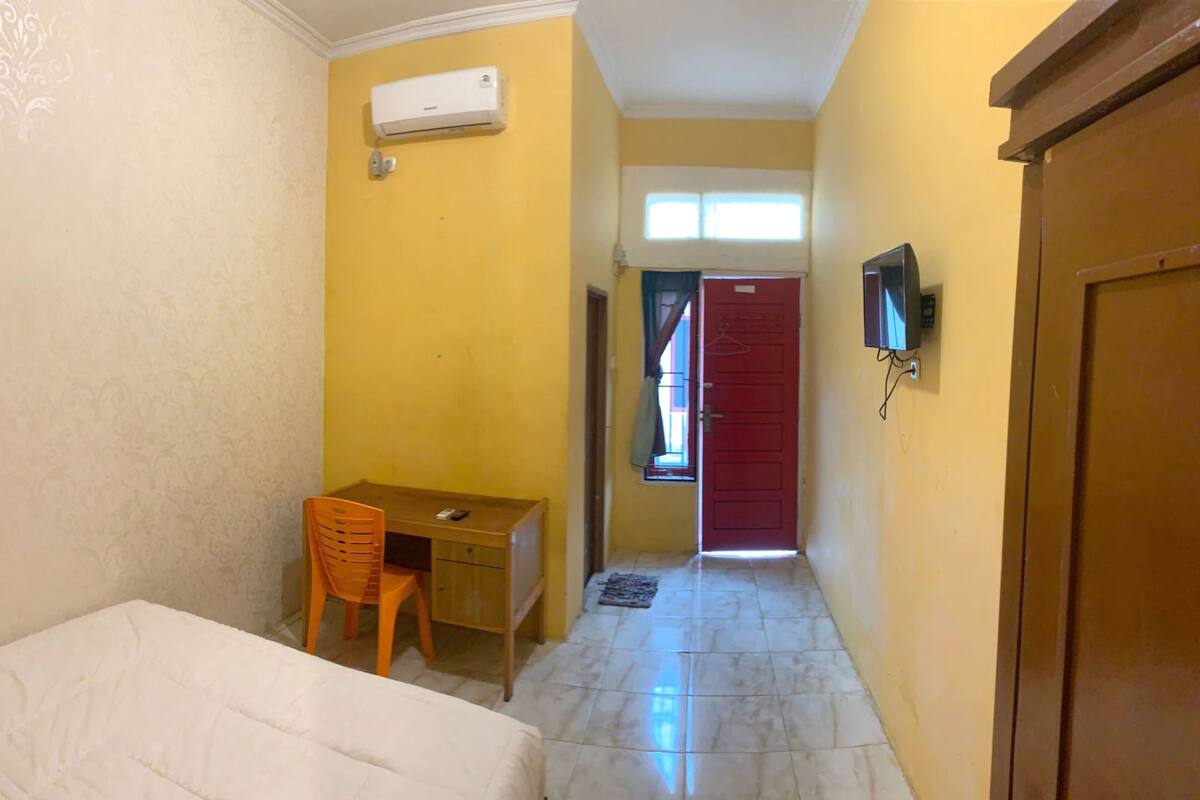 A hotel room with a bed, a desk with a chair, an air conditioning unit, a television, and a red door.