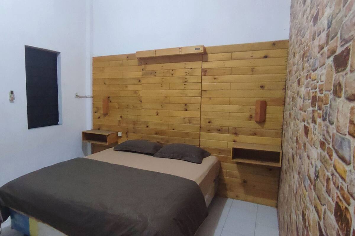 A bedroom with a wooden headboard, a bed with a brown bedspread, two windows, and a stone wall.