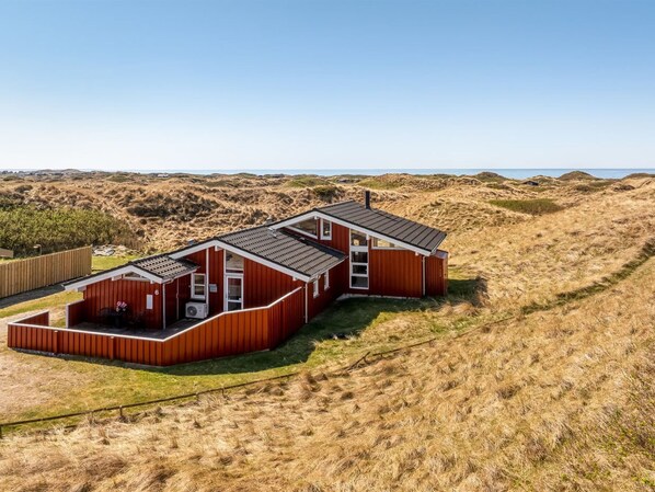 Una casa de madera roja con techo plano, un pequeño porche y vista a la playa y las dunas.