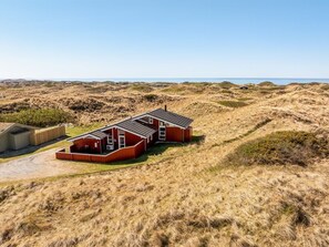A red wooden building with a flat roof, situated on a grassy dune with a clear sky above.