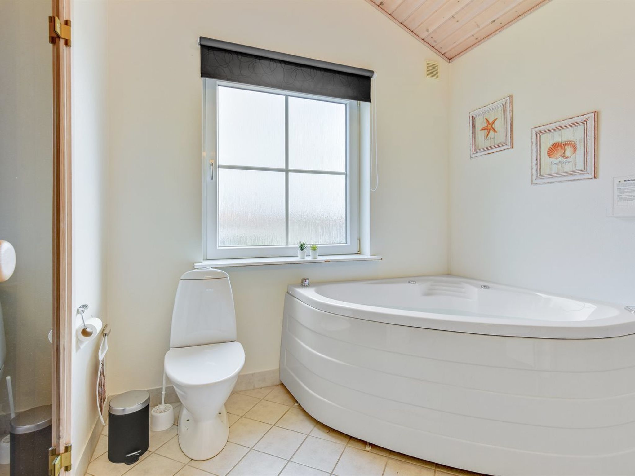 Ein Badezimmer mit Toilette, einem Fenster mit Jalousien, einer Badewanne und gerahmten Bildern an der Wand.