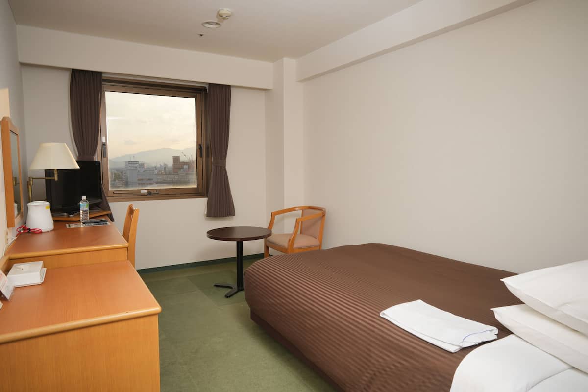 Une chambre d’hôtel avec un lit, un bureau, une chaise, une petite table, une lampe et une fenêtre avec des rideaux.