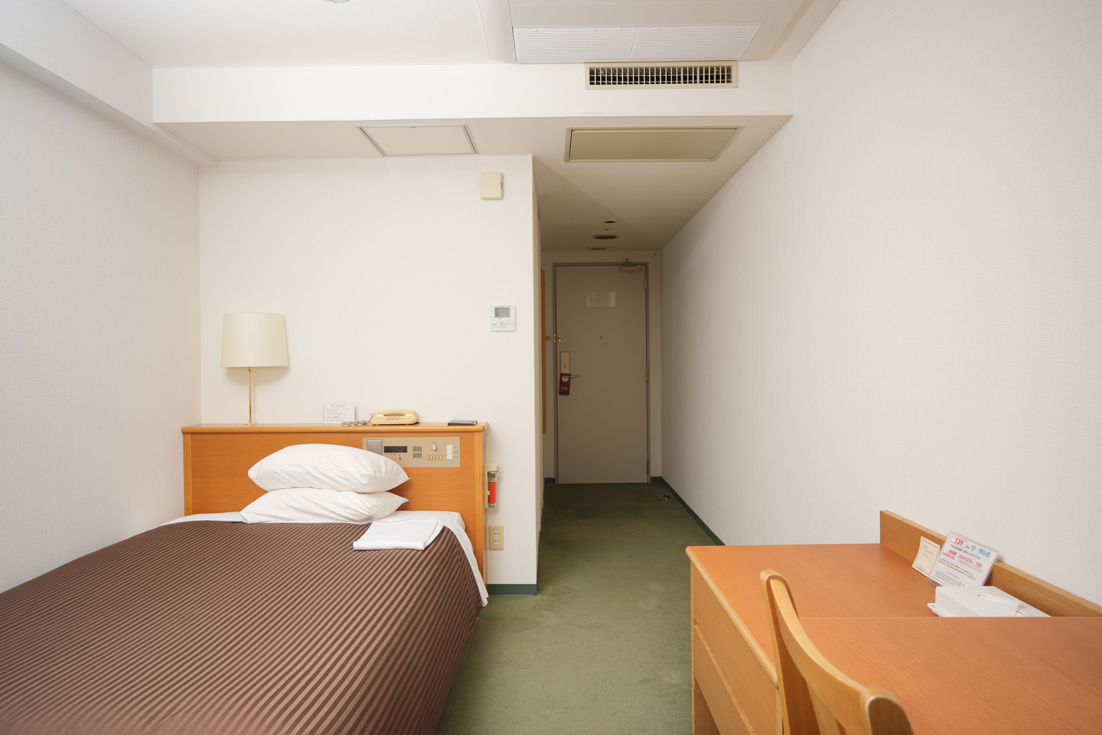 A hotel room with a single bed, a wooden desk, a lamp, and a small table with a sign.
