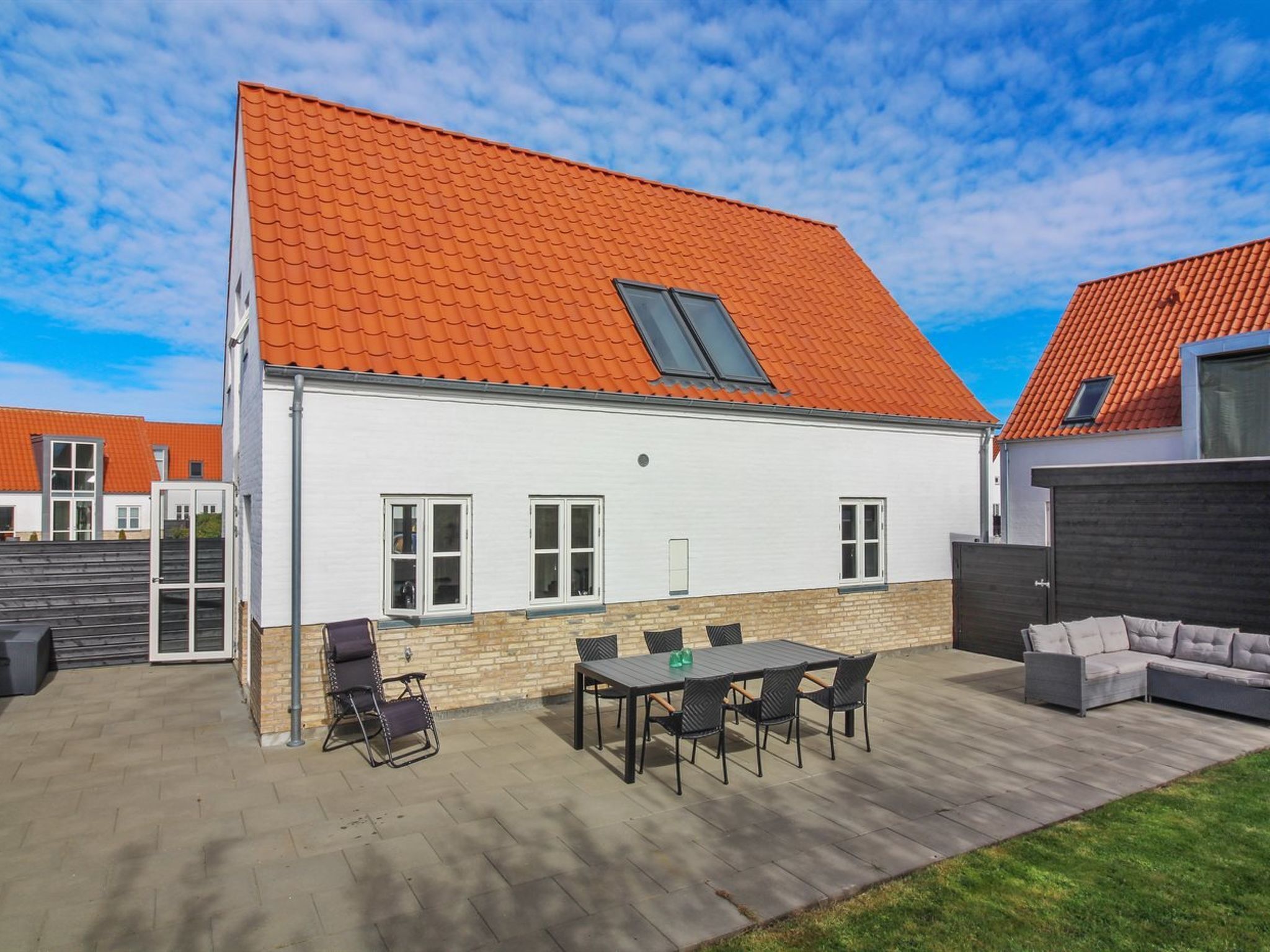 A modern house with a red-tiled roof, a patio with a table and chairs, and a grey sofa.
