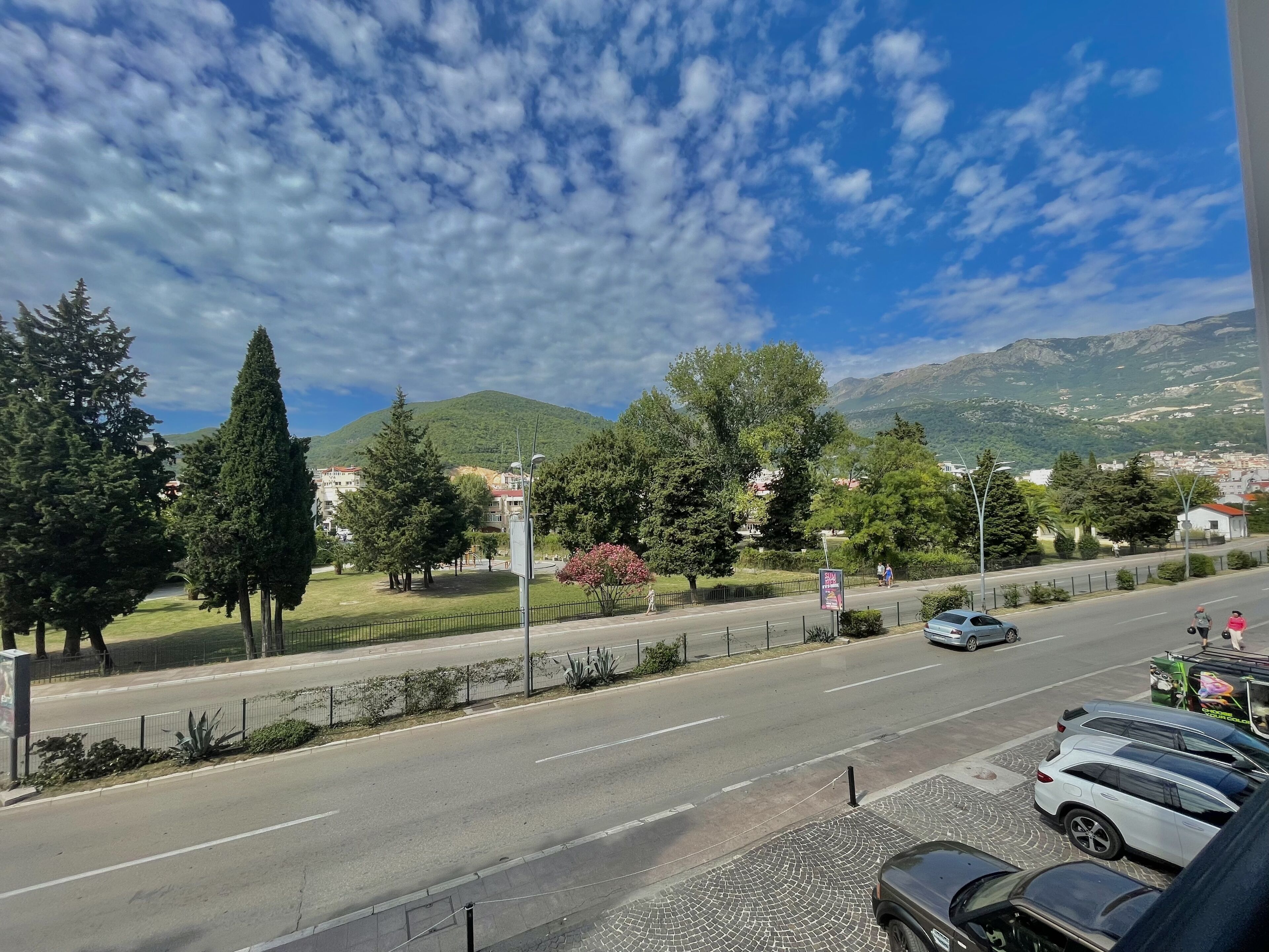 A street view with a road, parked cars, trees, and a mountainous backdrop.