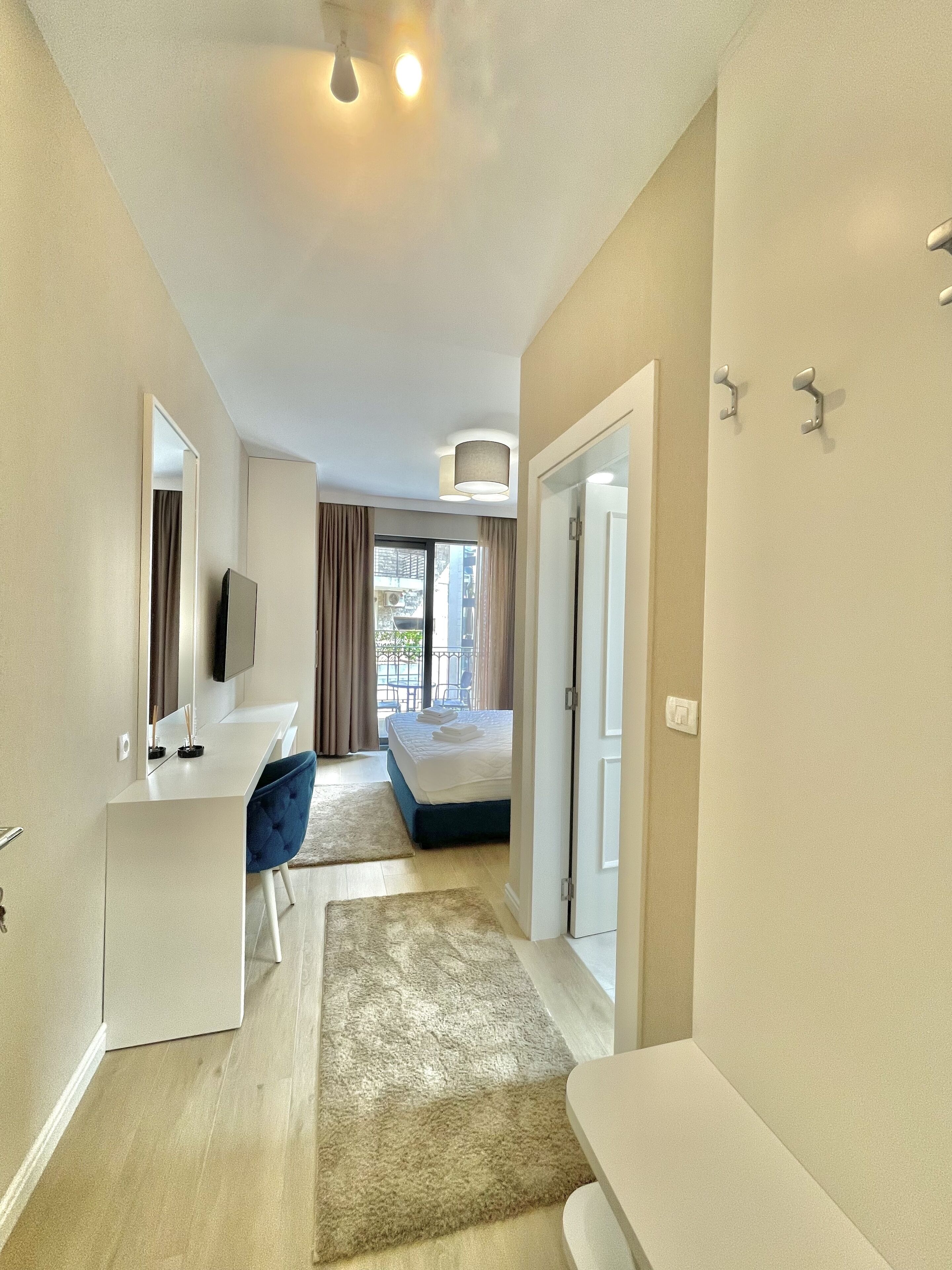 A modern hotel room with a bed, a desk, a chair, a TV, and a balcony with curtains.