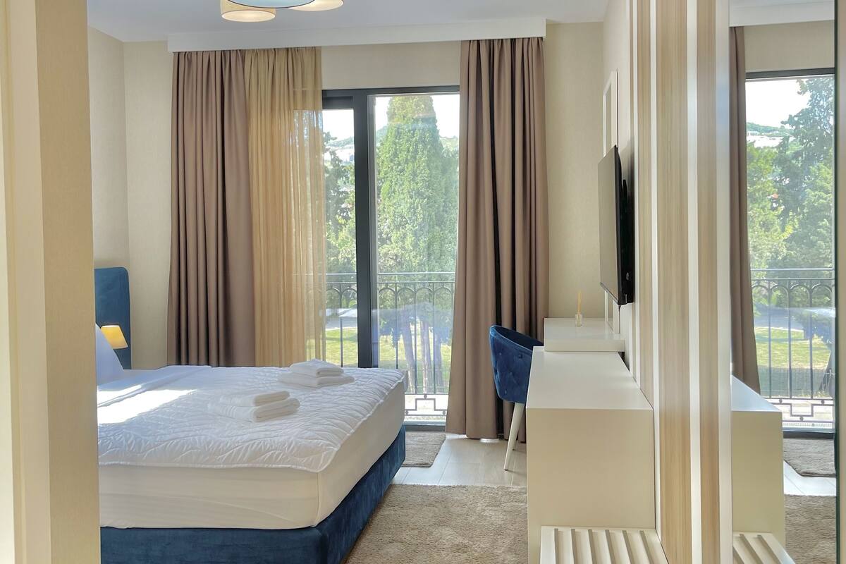A modern hotel room with a large bed, a desk, a TV, and a balcony with a view of greenery.