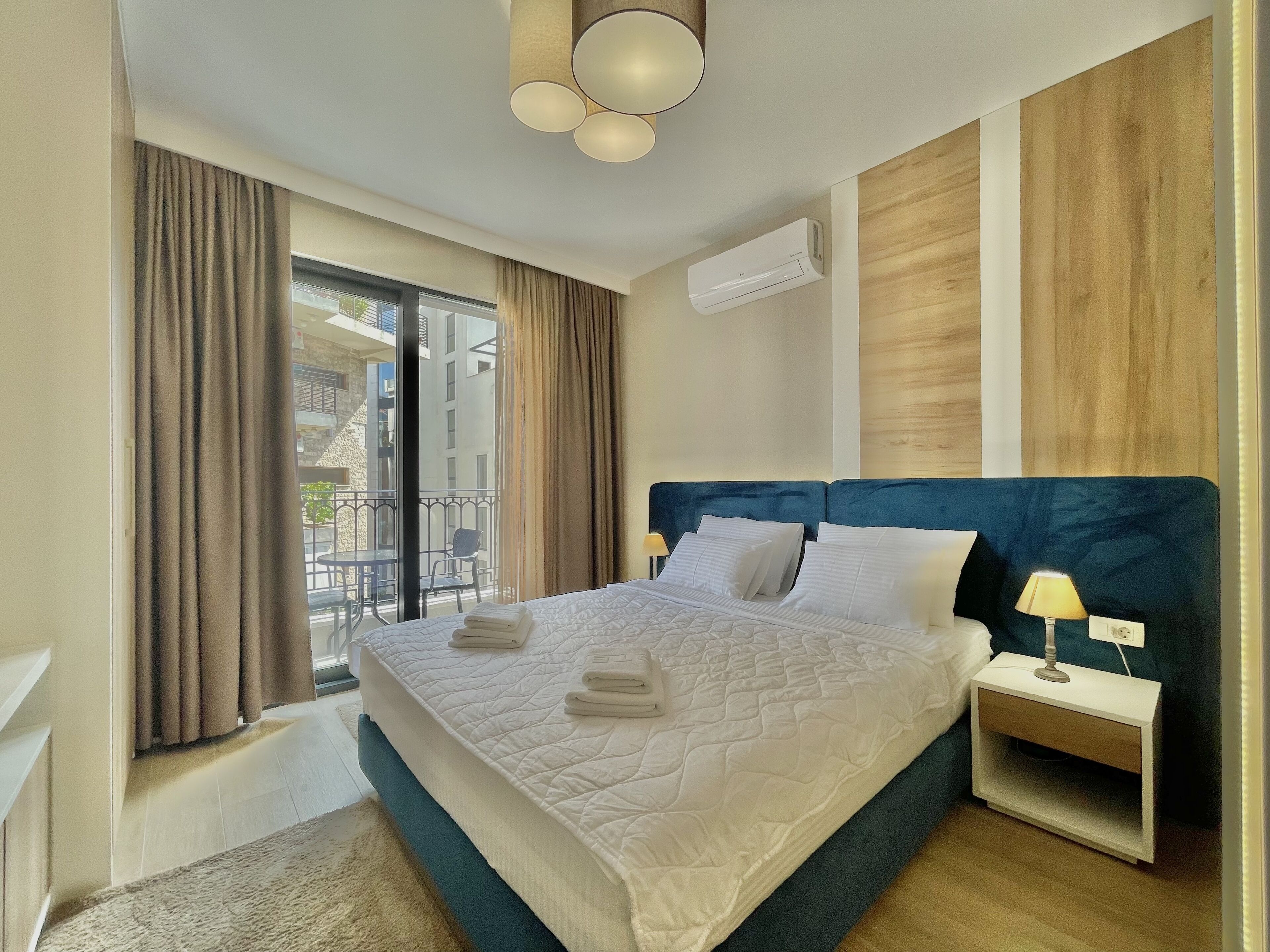 Habitación de hotel moderna con una cama grande, una mesita de noche, una lámpara, un equipo de aire acondicionado y vistas al balcón exterior.