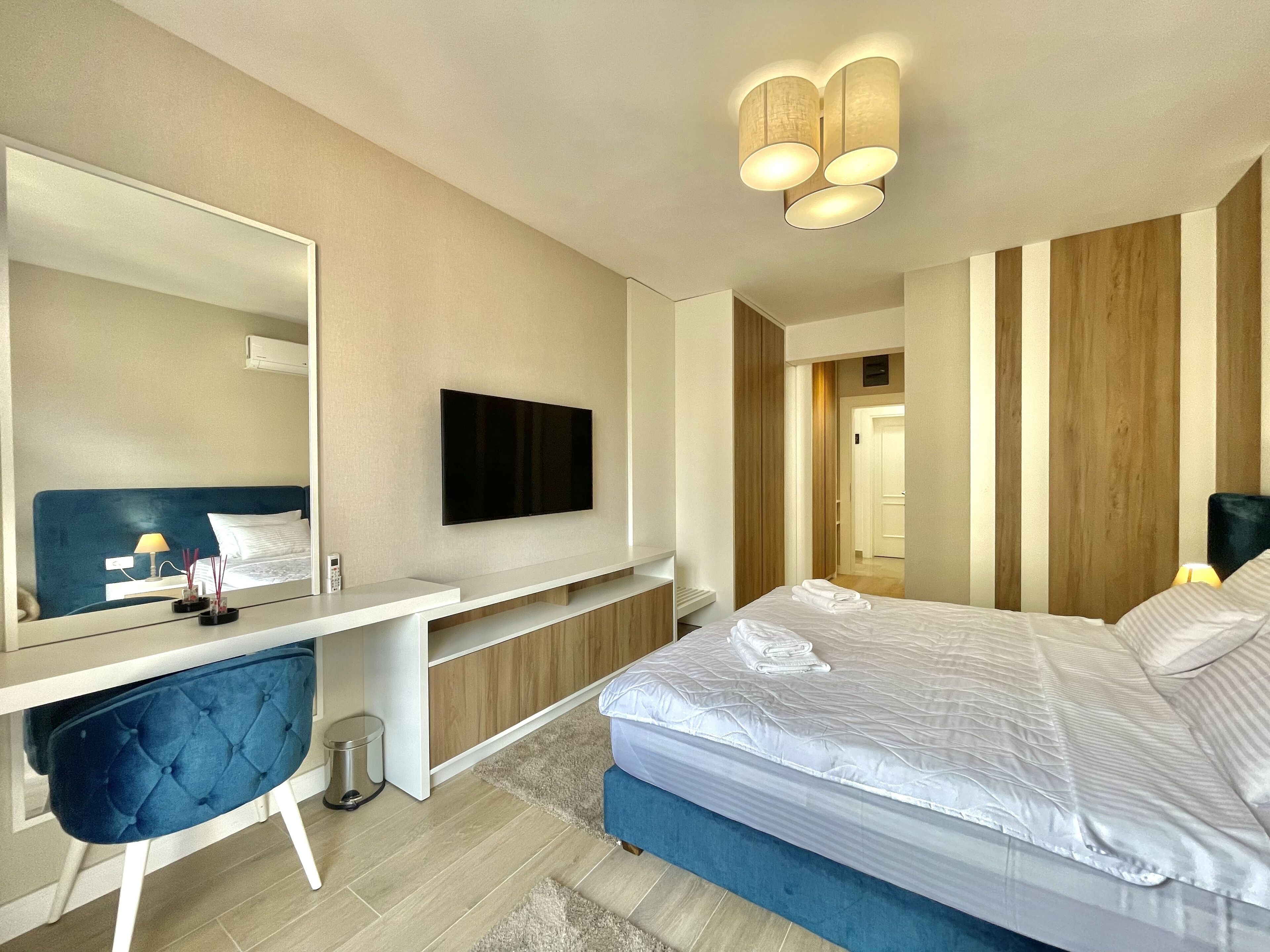 A modern hotel room with a large bed, a flat-screen TV mounted on the wall, a desk with a chair, and a mirror.