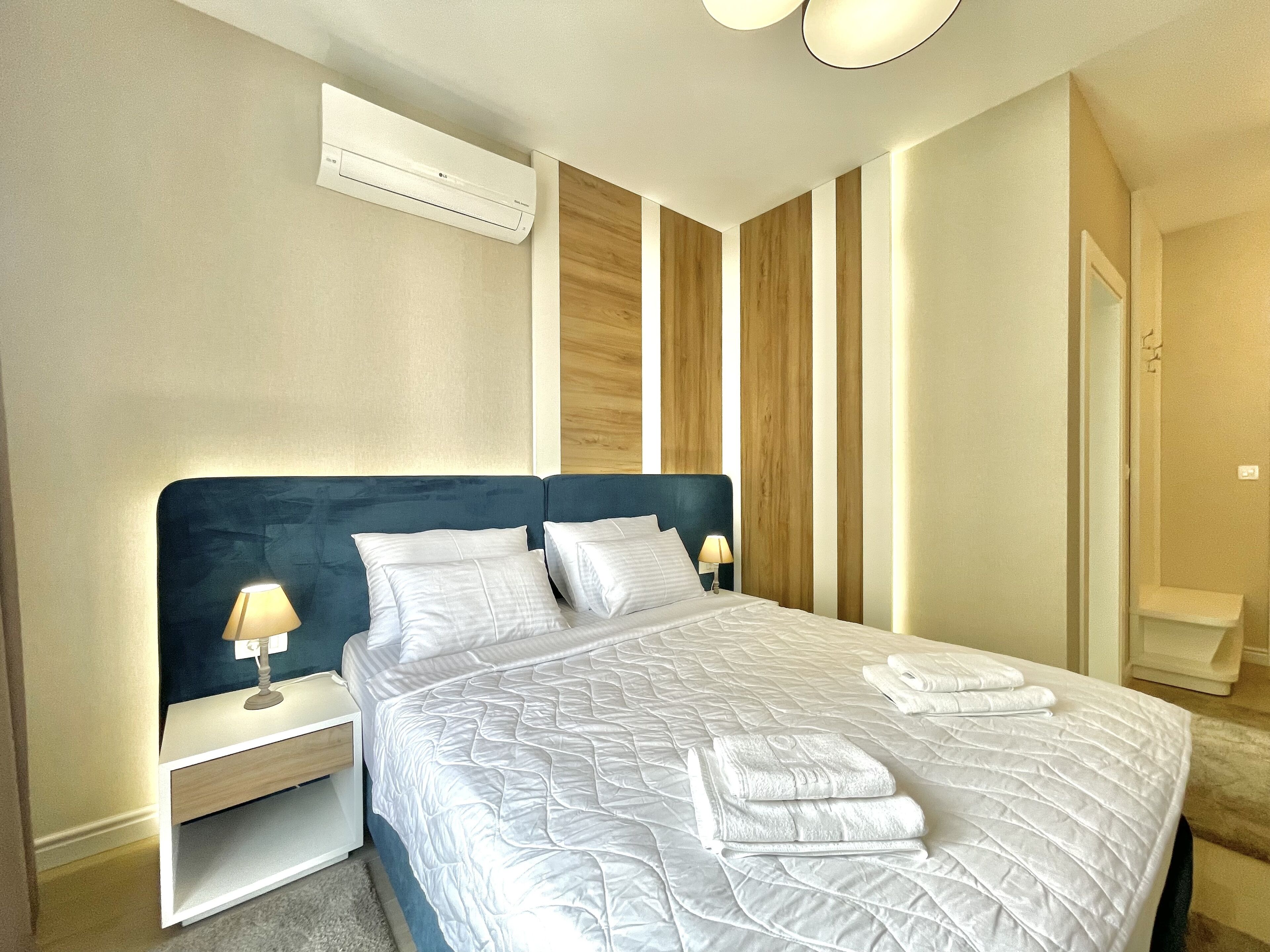 A neatly made bed with white linens and towels, a bedside lamp, and a wall-mounted air conditioner.