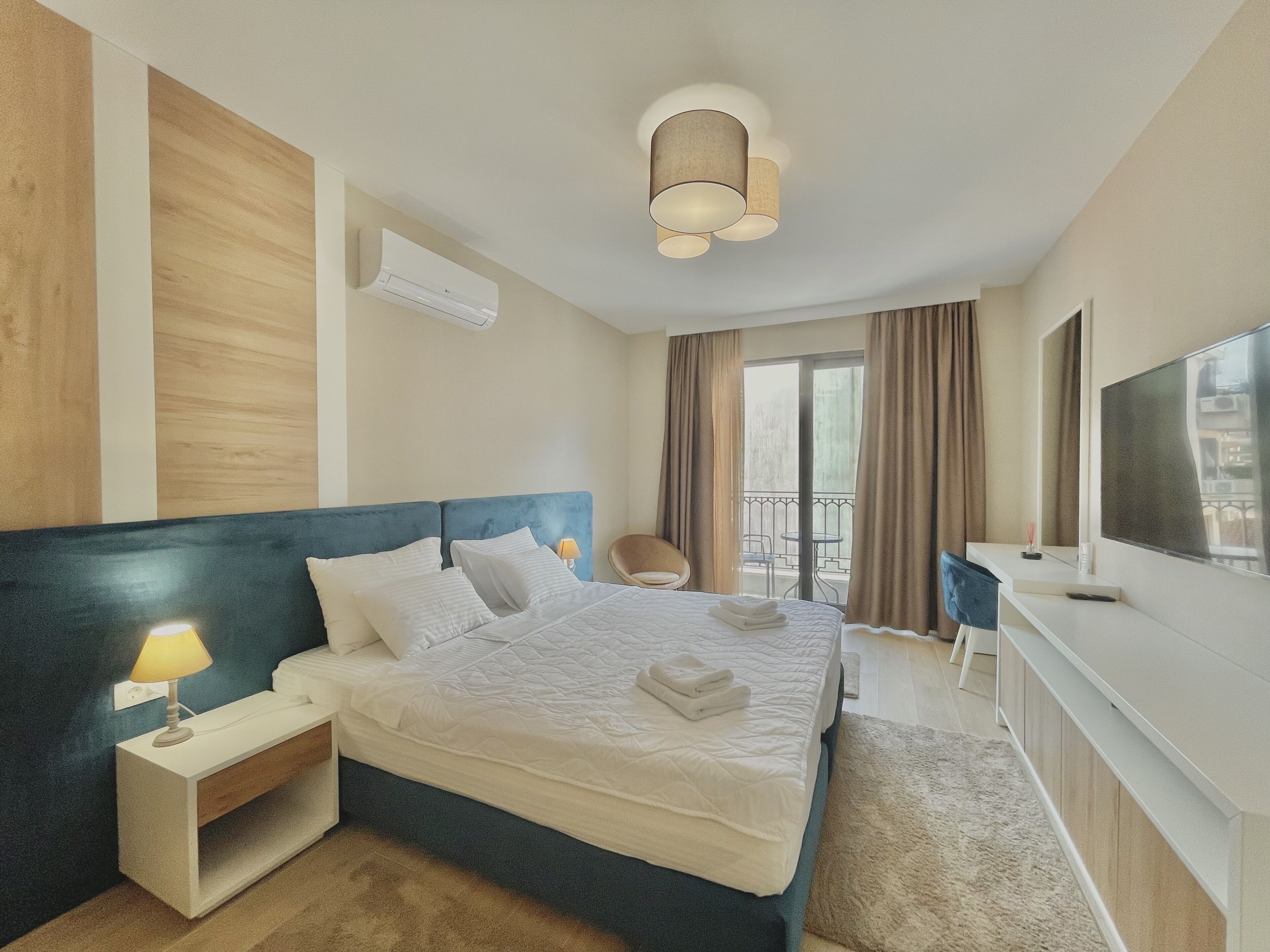Une chambre d’hôtel avec un grand lit, une table de chevet avec une lampe, un bureau avec une chaise, une télévision et un balcon avec des rideaux.