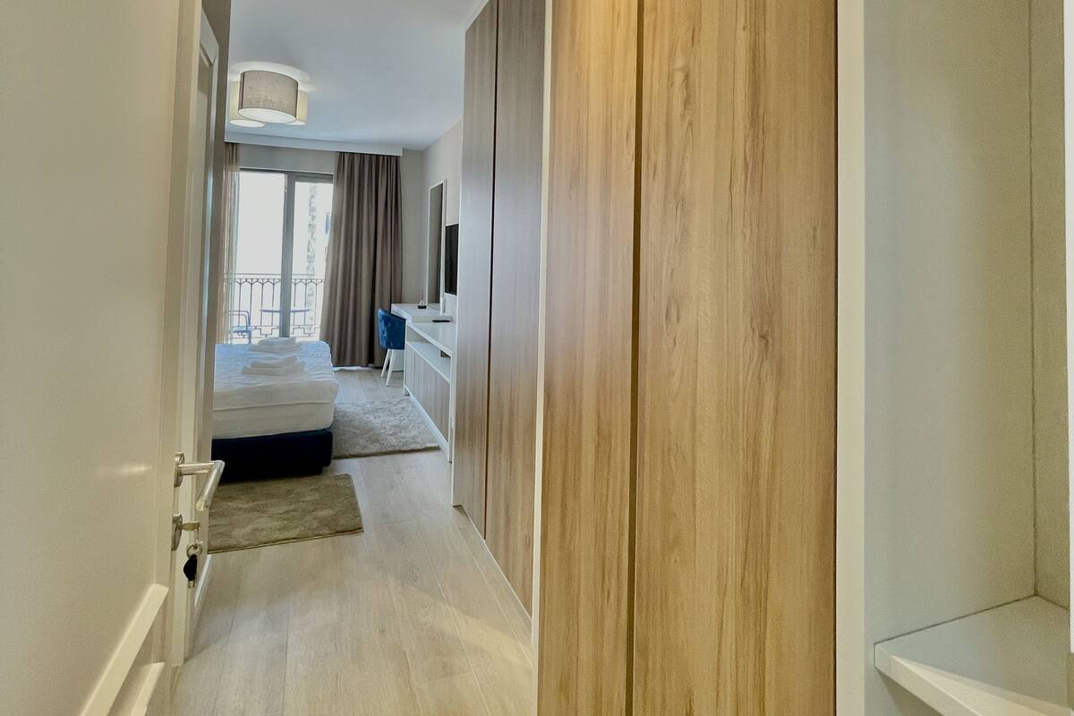 A modern hotel room with a wooden wardrobe, a bed with a white duvet, a desk with a chair, and a view of the balcony.