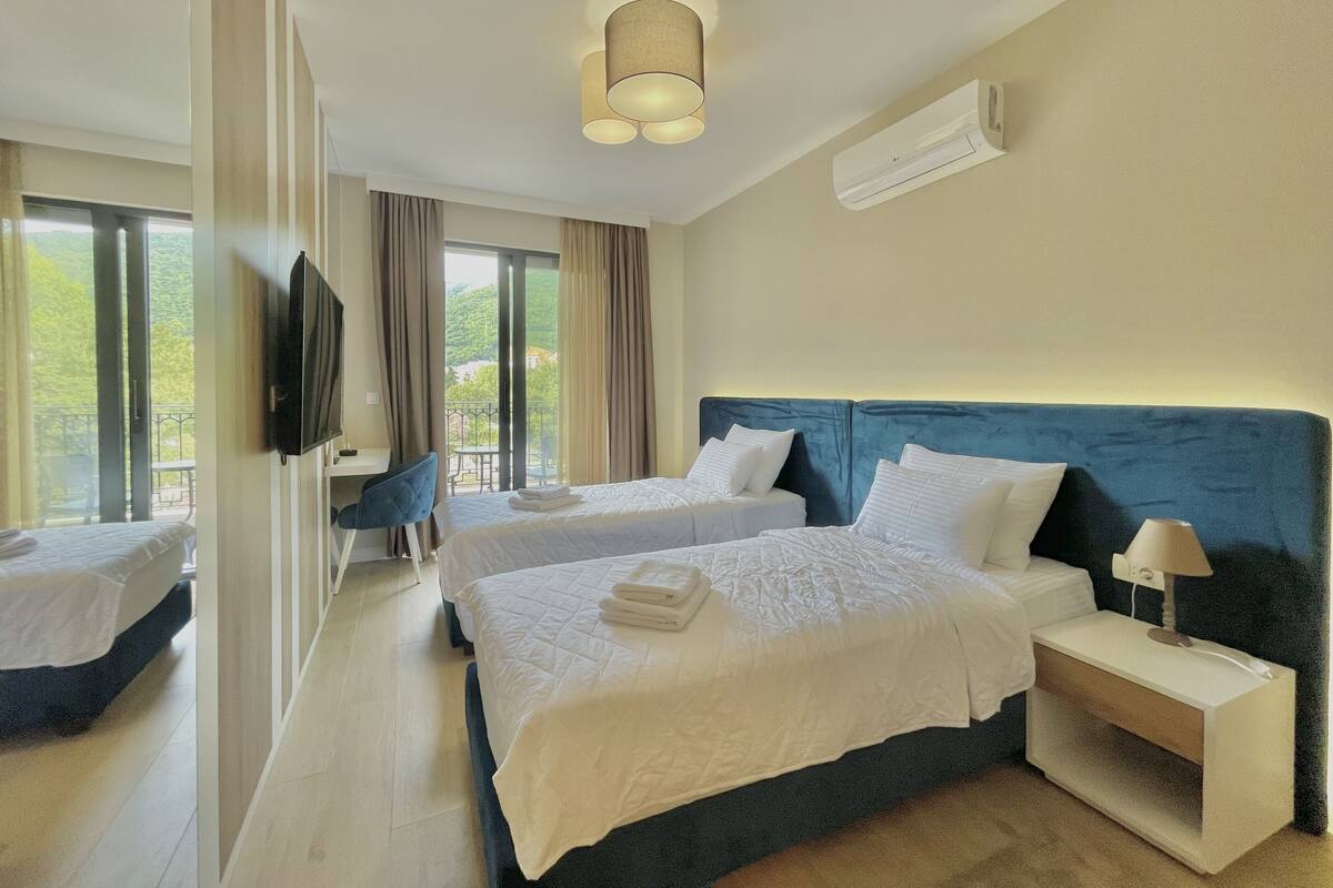 A hotel room with two beds, a TV, a desk, and a balcony with a view.