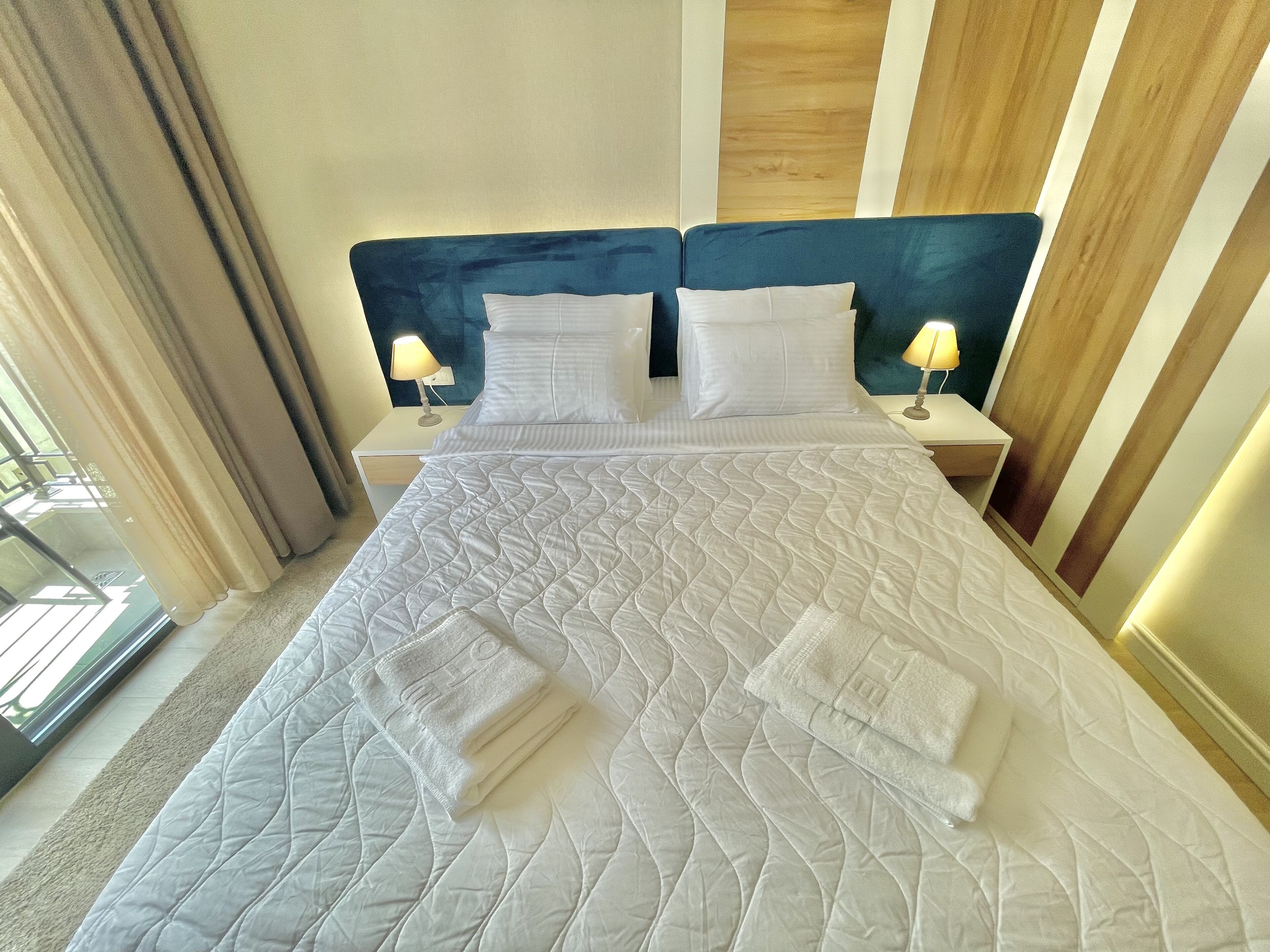 Una habitación de hotel con una cama grande, dos mesitas de noche con lámparas y vistas al exterior a través de una ventana con cortinas.