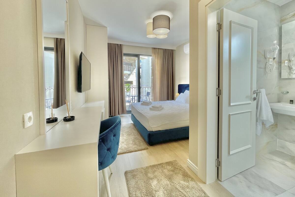 A hotel room with a bed, a desk, a chair, a bathroom with a sink and mirror, and a balcony with curtains.