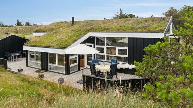 A modern house with a deck, large windows, and a grassy hill in the background.