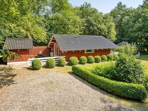A red wooden cabin with a sloped roof, surrounded by well-manicured hedges and a gravel driveway.