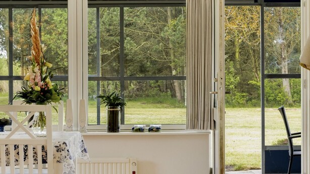 A room with a white radiator, a white chair, and a table with a floral tablecloth. There are two windows with curtains, one of which is open, and a view of a garden outside. - "Mikkeline" - 400m to the fjord by Interhome (Øster Assels)