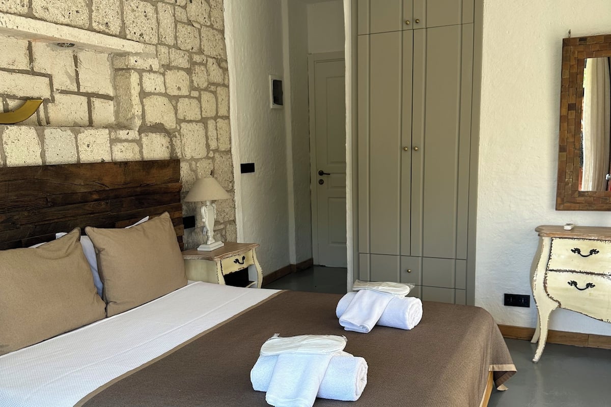 A bedroom with a stone wall, a bed with beige and brown bedding, a nightstand with a lamp, and a wardrobe.
