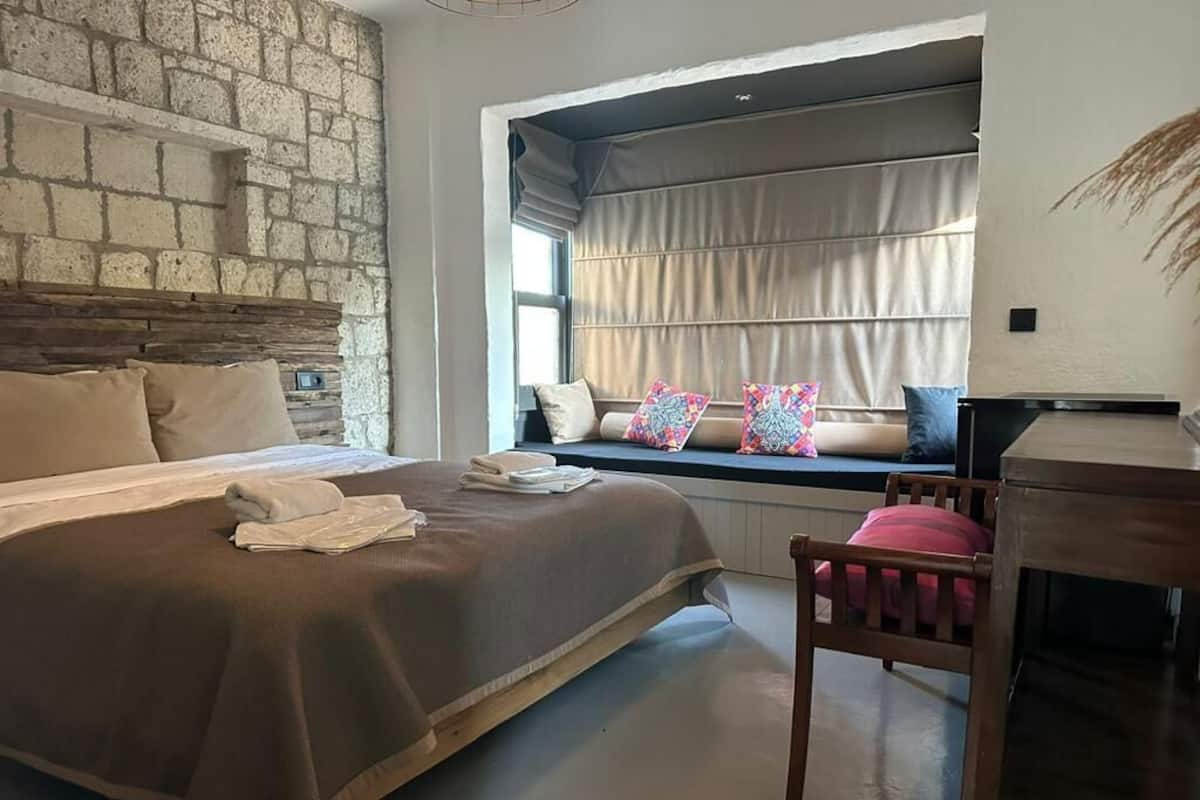 A bedroom with a large bed, a stone wall, a window with curtains, and a wooden chair.