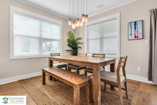 Un coin repas avec une table et des chaises en bois, de grandes fenêtres équipées de stores, un luminaire suspendu et une plante en pot.