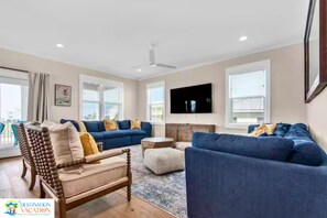 A modern living room with a blue sofa set, a wooden chair, a flat-screen TV mounted on the wall, and a ceiling fan. - Beachside Escape w/ Pool, Views & Pickleball Fun (Gulf Shores)