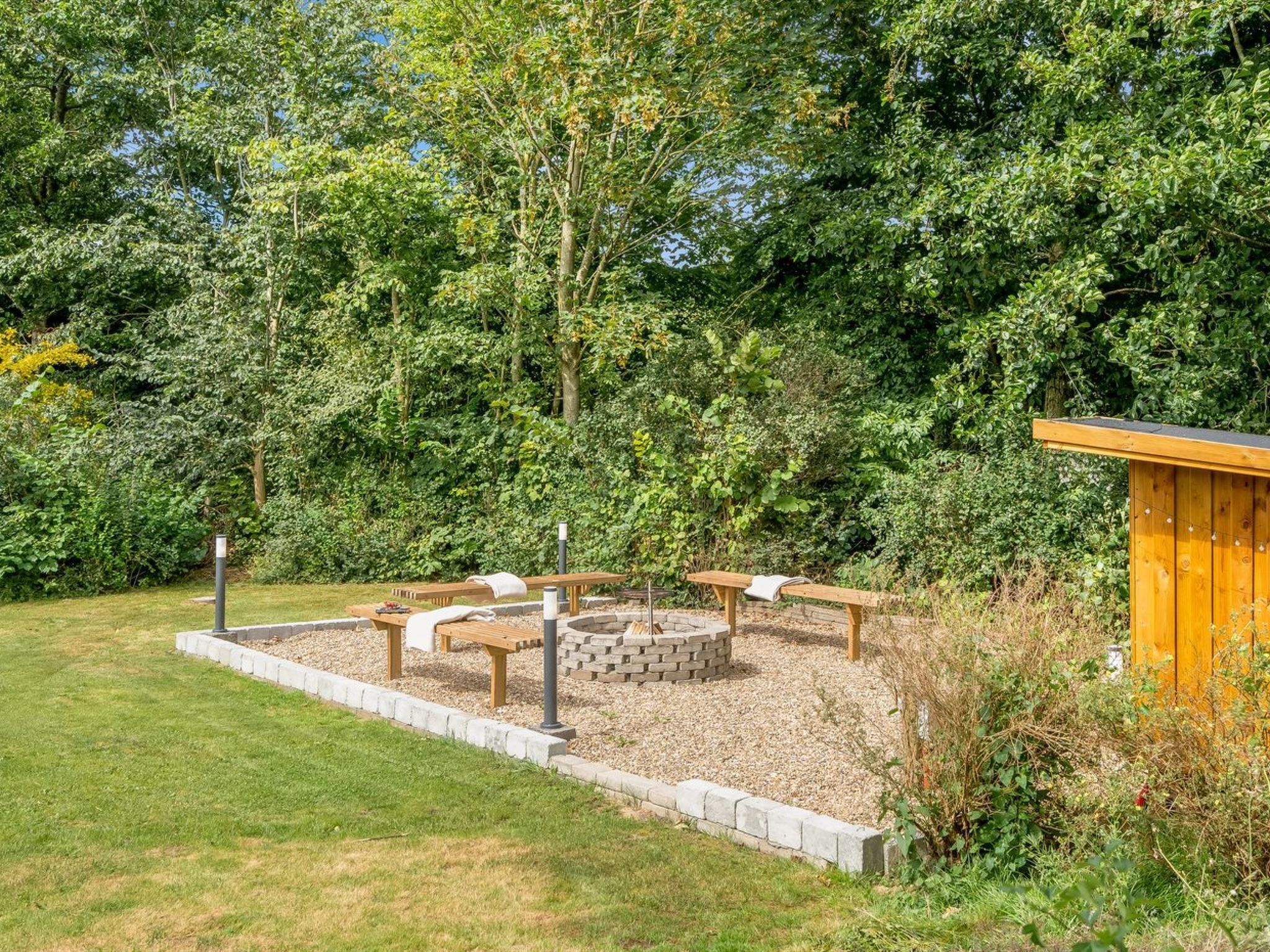 Un giardino con un braciere, panche di legno e una piccola costruzione in legno.