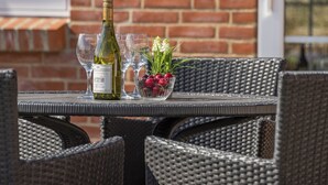 Een tafel met een fles wijn, glazen en een schaal fruit, geplaatst bij een rotan stoel.