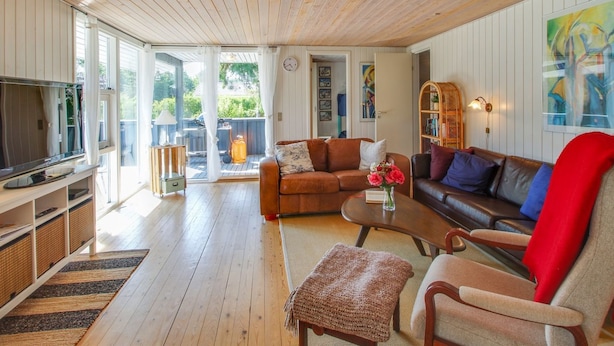 Un salon chaleureux avec un plafond en bois, un canapé en cuir marron, un fauteuil rouge, une table basse ornée d’un vase de fleurs et un téléviseur posé sur un meuble.