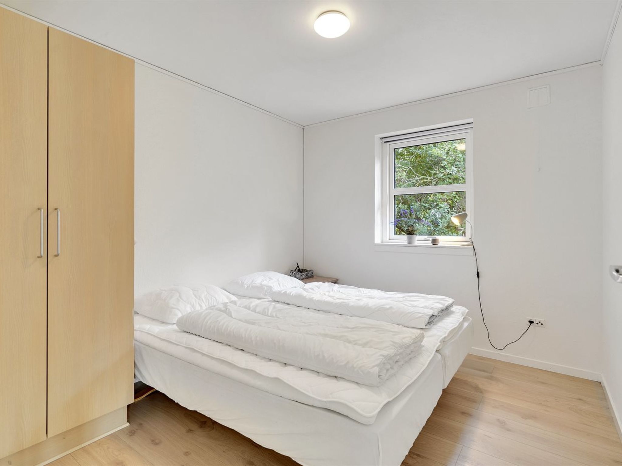 Un dormitorio con cama, un armario de madera, una ventana con vistas a zonas verdes y un enchufe en la pared.