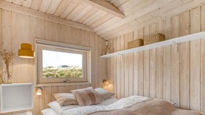 Un dormitorio con un ventanal grande, paredes de madera, una cama con ropa de cama blanca, un plaid tejido y una estantería con cestas.