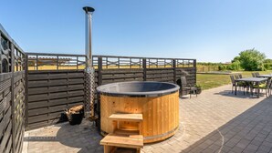 Un bain à remous avec une structure extérieure en bois et une cheminée en métal, entouré d’une terrasse en bois et de mobilier d’extérieur.