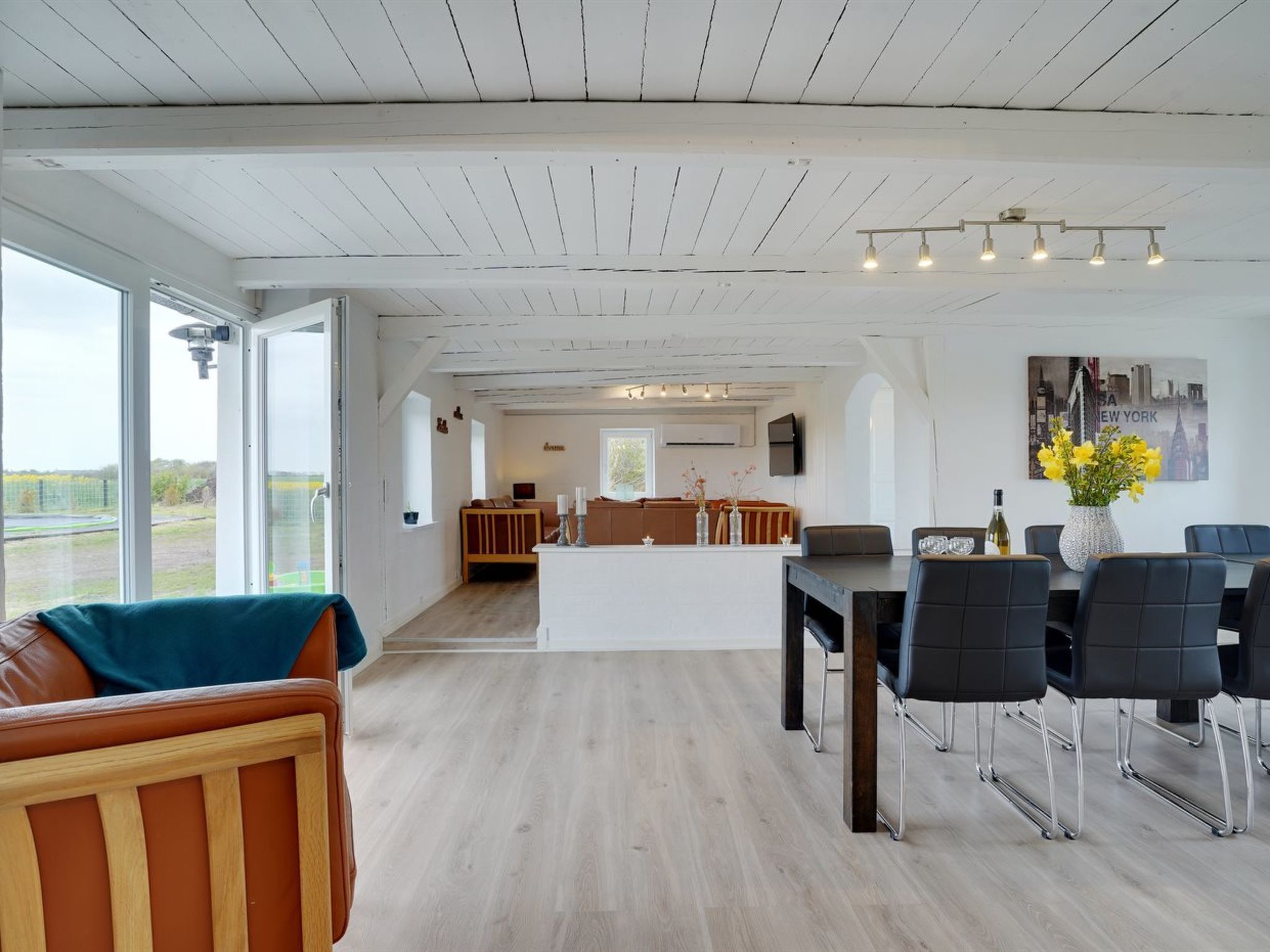 Een ruime woonkamer met een houten plafond, een eettafel met stoelen, een bank en een keuken op de achtergrond.