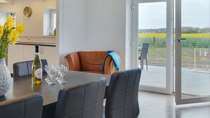 Un coin repas avec une table dressée pour quatre personnes, un fauteuil en cuir marron et une vue sur un champ jaune, visible à travers une porte vitrée.