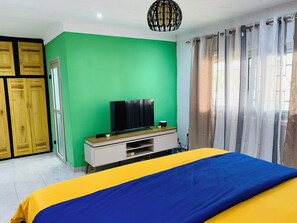 Ein Schlafzimmer mit grüner Wand, einem Bett mit gelb-blauer Bettdecke, einem Fernseher auf einem Holzschrank und einem Fenster mit Vorhängen.