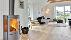 Un salon moderne avec un poêle à bois, un canapé, un fauteuil et une vue sur l’extérieur.