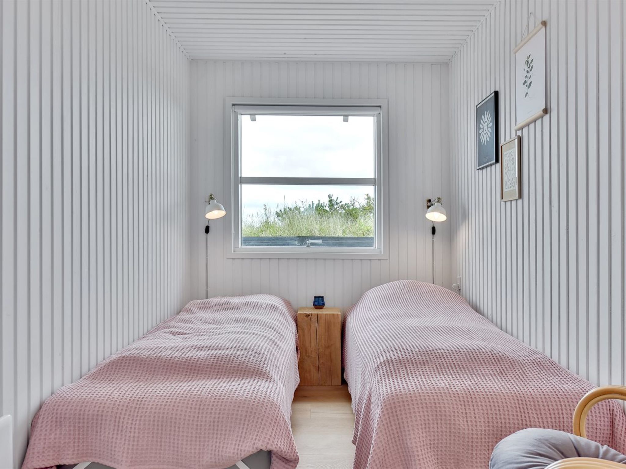 A room with two beds, a window with a view of grass, and wall-mounted lamps.