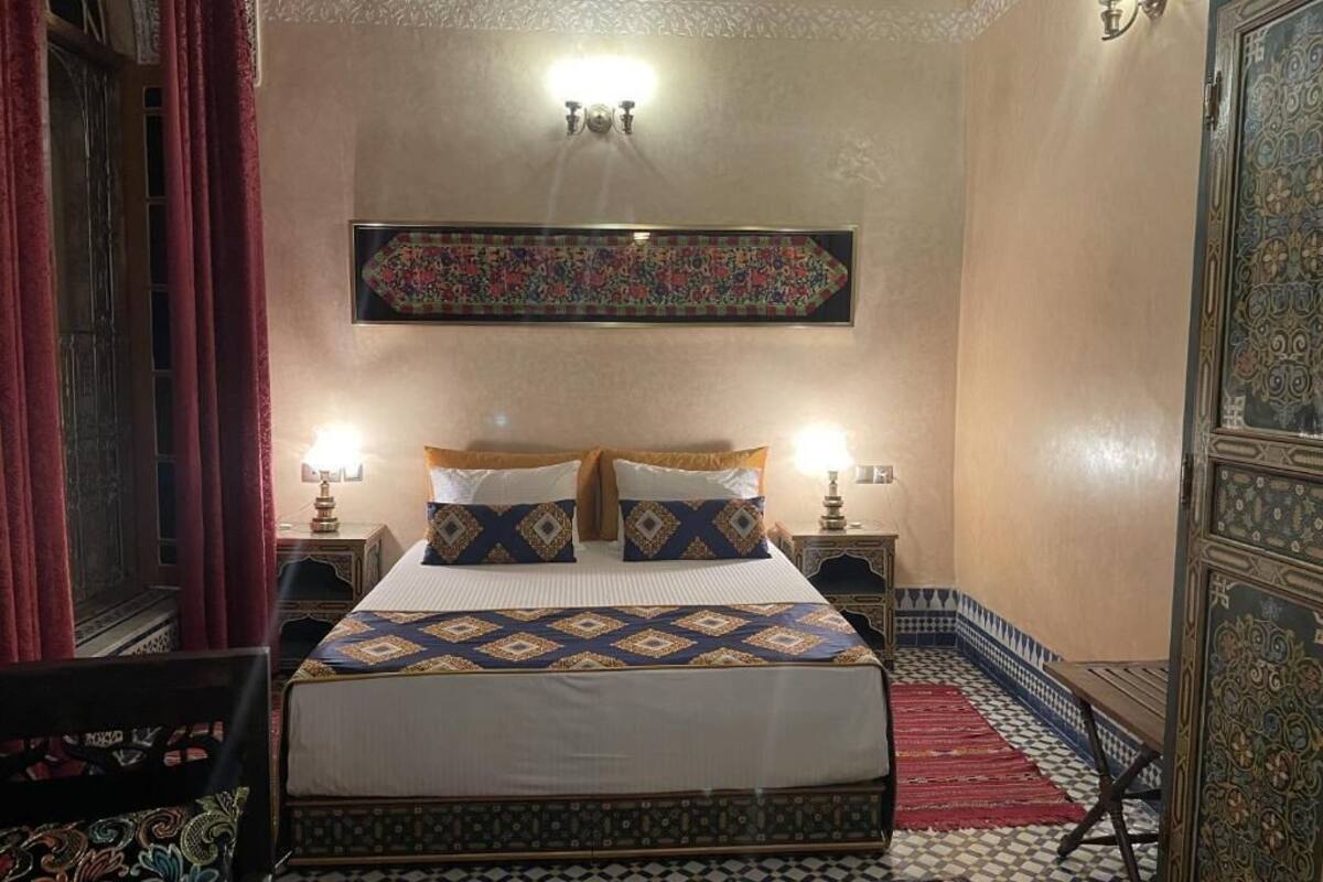 A traditional Moroccan room with a bed, two bedside lamps, a patterned tapestry, and a decorative door.