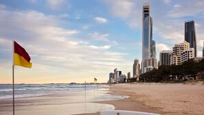 A beach with a lifeguard stand and a surfboard, a flag, and a city skyline in the background. - Perfect 4-bed Penthouse w/ Parking, Pool and Gym (Melbourne)