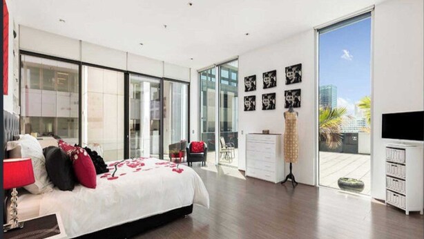 A modern bedroom with a large bed, a dresser, a TV, and a balcony with a city view. - Perfect 4-bed Penthouse w/ Parking, Pool and Gym (Melbourne)