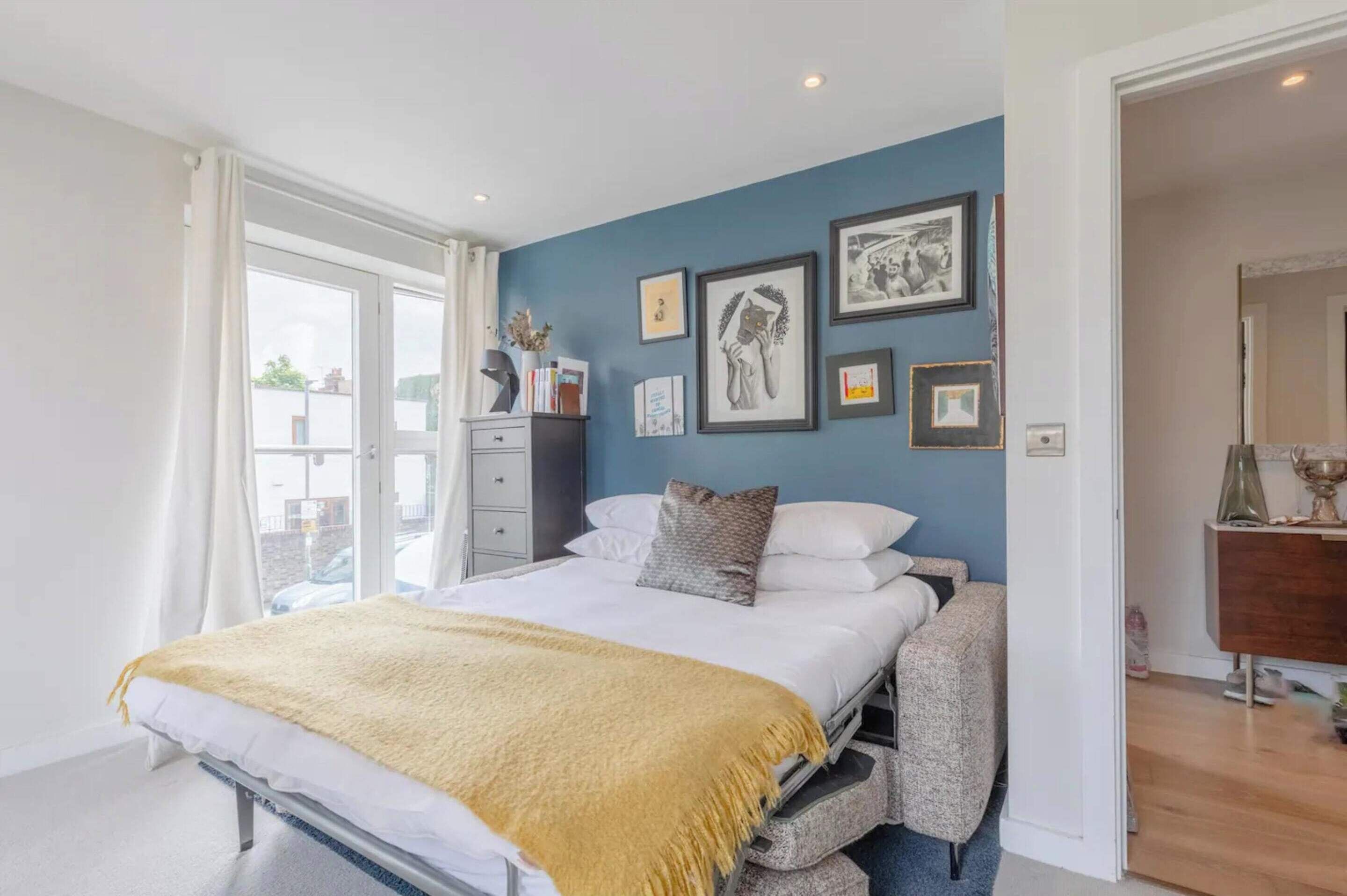 A bedroom with a bed, bedside table, and a dresser. There are framed pictures on the wall and a window with curtains.