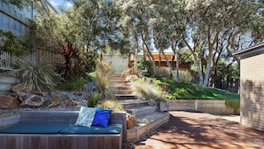 A wooden deck with a built-in bench and cushions, surrounded by a landscaped garden with various plants and a wooden staircase leading to a building.