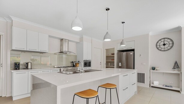 A modern kitchen with white cabinetry, a central island with stools, stainless steel appliances, pendant lighting, and a wall clock.