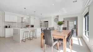 A modern kitchen with white cabinets, a central island with stools, a dining table with chairs, and a large window providing natural light.