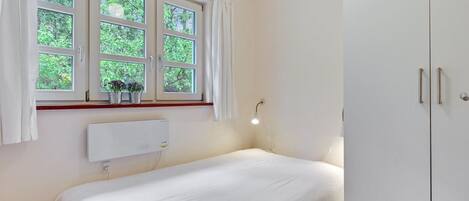 A bedroom with a bed, a window with curtains, a bedside table with a lamp, and a wardrobe.