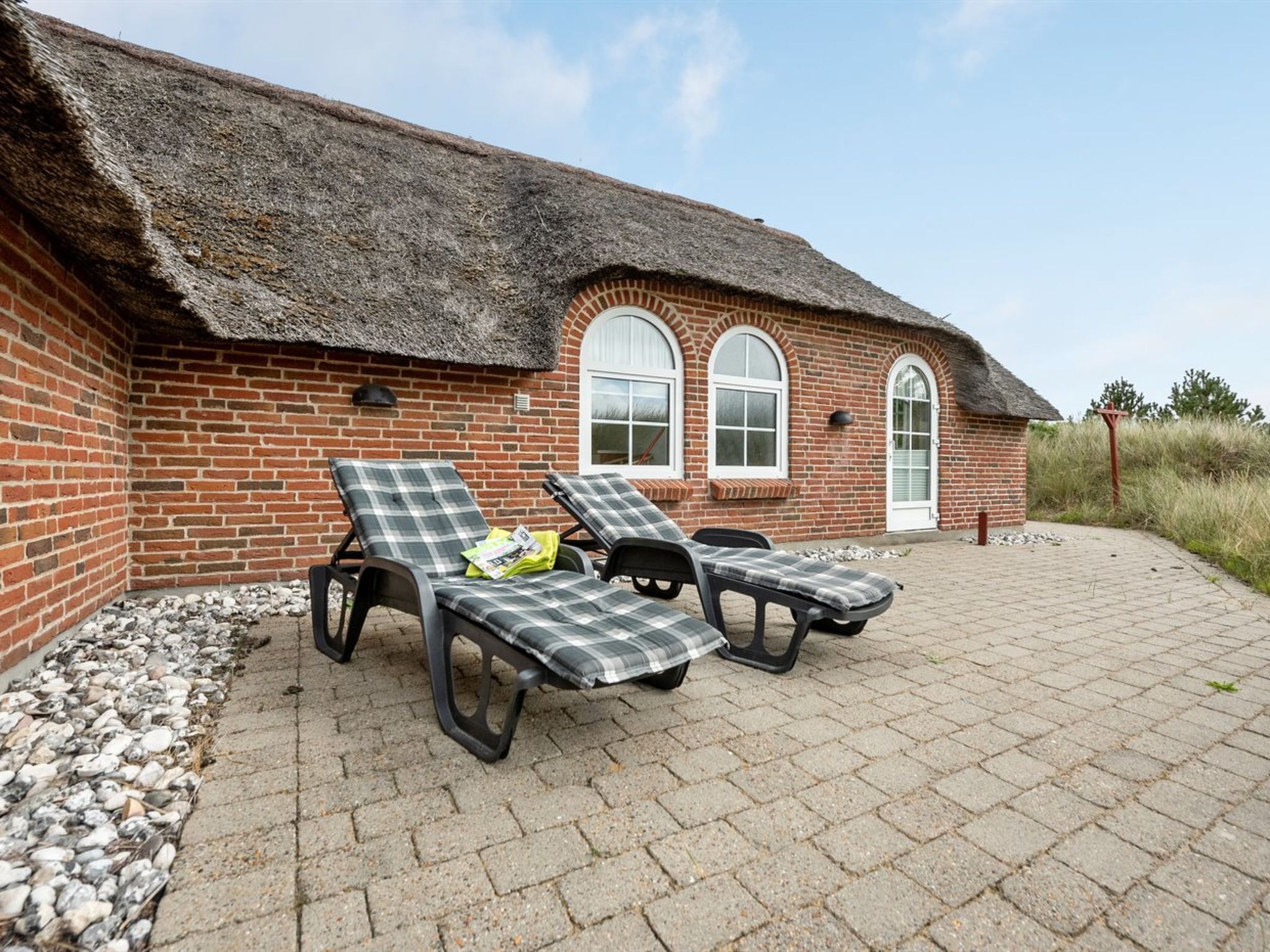 A brick building with a thatched roof, two lounge chairs with checkered cushions, and a paved patio area.