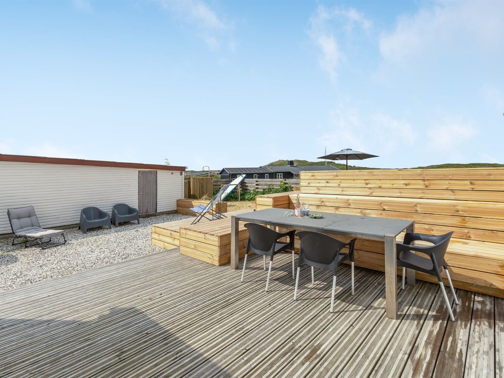 Eine Holzterrasse mit Tisch und Stühlen, einem Grill und einem überdachten Bereich mit Sonnenschirm.
