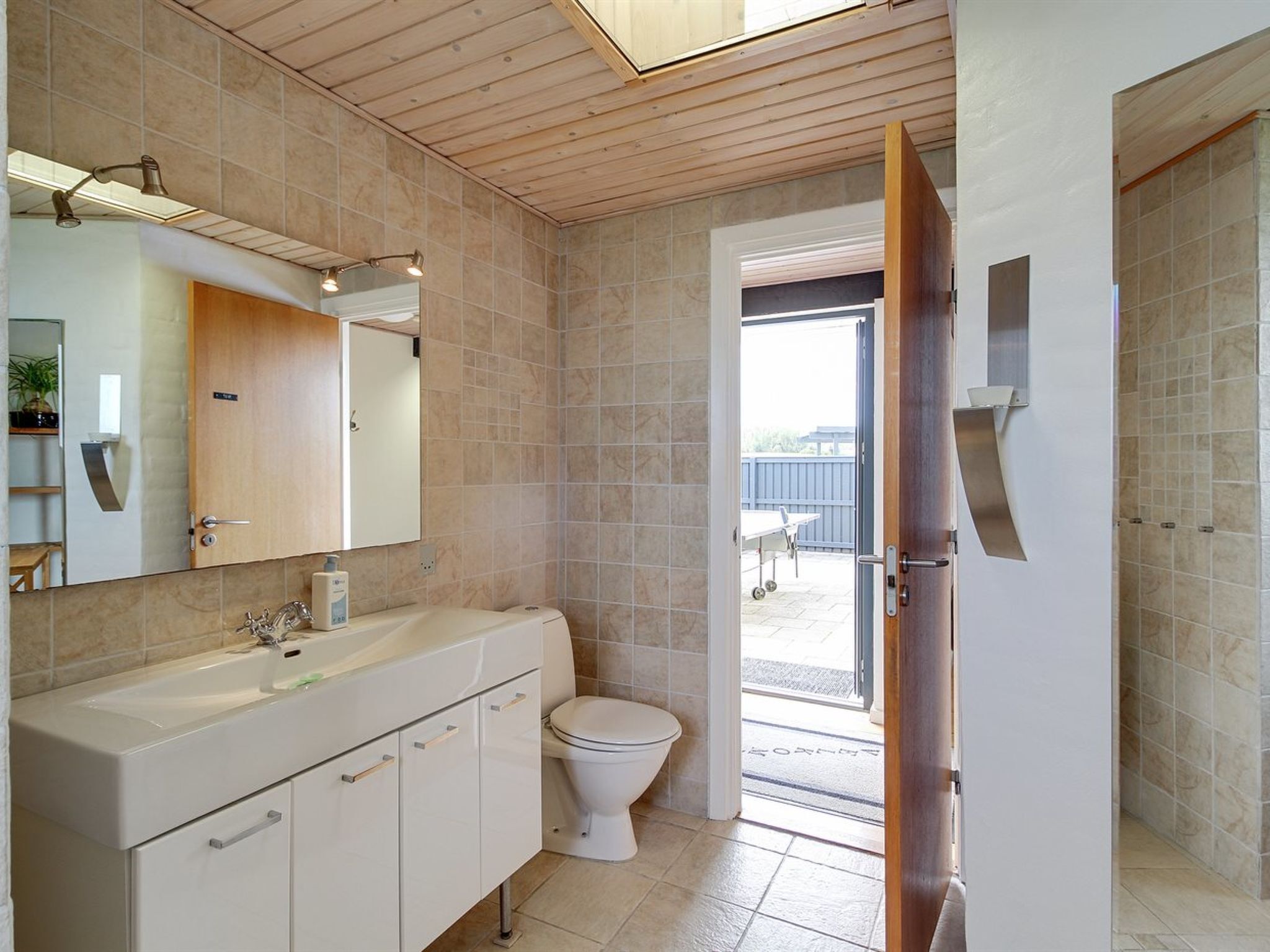 A modern bathroom with a wooden ceiling, a large mirror, a white sink, and a toilet.