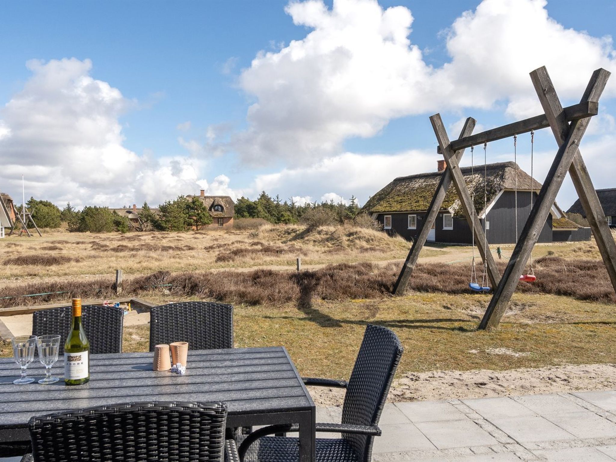 Un espace extérieur aménagé avec une table et des chaises, une bouteille de vin, et un portique à balançoire en arrière-plan.
