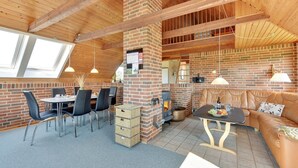 Ein gemütlicher Innenraum mit Kamin aus Backstein, Holzbalken, einer Essecke mit Stühlen, einem Sofa und einem Tisch mit einer Fruchtschale.
